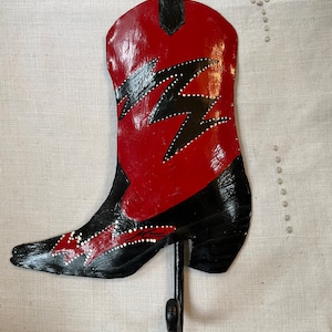 May include: A red and black cowboy boot shaped wall hook with a lightning bolt design. The hook is made of metal and has a black finish.