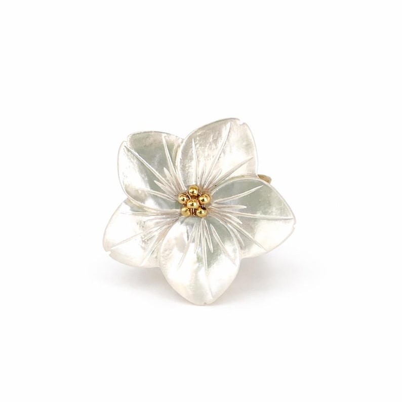 May include: A flower-shaped ring with five white petals and a cluster of small gold beads at the center. The ring is set against a white background, highlighting the delicate details of the floral design.