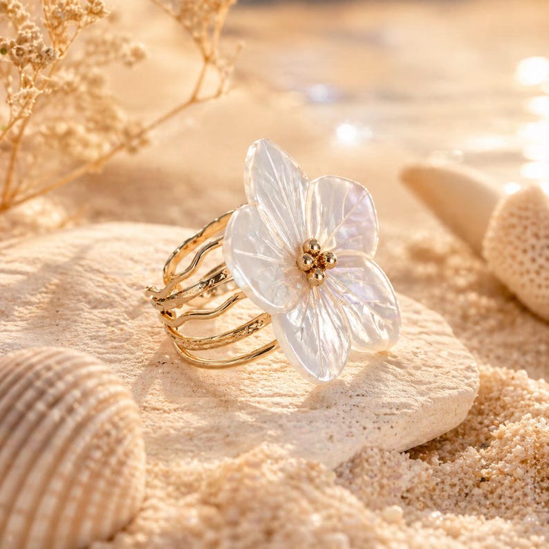 Peut inclure: Une bague dorée avec une grande pièce maîtresse en forme de fleur blanche. La fleur a des pétales délicats et translucides et un groupe de petites perles dorées en son centre. La bague est posée sur une pierre claire, avec un fond sablonneux.