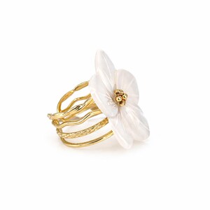 May include: A gold-toned ring featuring a white flower design. The ring has multiple bands with a textured pattern. The flower has a cluster of gold-colored beads at its center. The ring is set against a white background.