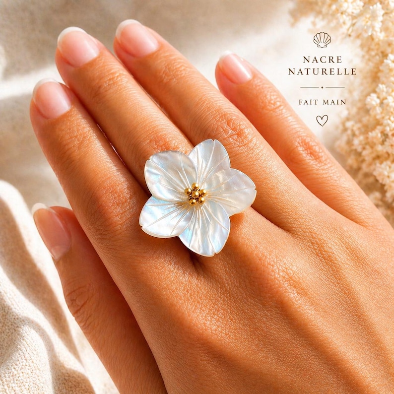 May include: A white flower-shaped ring with iridescent petals and a gold center is displayed on a hand. The ring's design resembles a five-petal flower. The ring is a piece of jewelry.