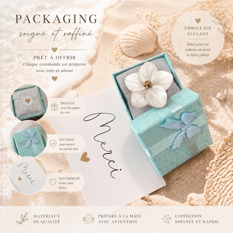 May include: A light blue gift box tied with a blue ribbon, containing a white flower with a gold center. The box sits on a sandy surface next to a seashell and a card that says "Merci". The packaging is described as "soign&eacute; et raffin&eacute;".