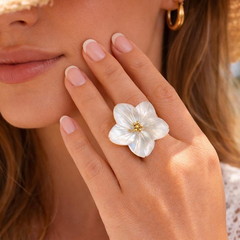 May include: A white flower-shaped ring with a gold center, worn on a finger. The ring's petals have a pearlescent sheen. The jewelry is a floral design, perfect for adding a touch of nature-inspired elegance to any outfit.
