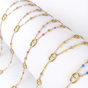 Op de afbeelding: Goudkleurige kettingen met ovale hanger en kleurrijke kraalaccenten. De kettingen hebben roze, gele en blauwe kralen. De kettingen zijn over een wit oppervlak gedrapeerd. De sieraden zijn geschikt voor dagelijks gebruik.