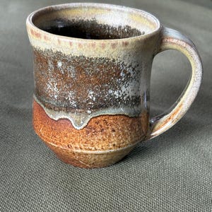 Könnte beinhalten: Eine handgefertigte Keramiktasse mit rundem Boden und gebogenem Henkel. Die Tasse hat eine strukturierte Oberfläche mit einer Mischung aus Braun-, Beige- und Orangetönen. Das Innere ist dunkel, und der Rand hat ein helleres, gesprenkeltes Aussehen.