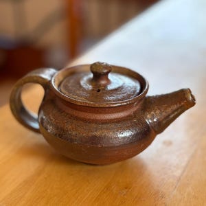 May include: A handcrafted, brown ceramic teapot with a rounded body, a curved handle, and a spout. The lid has a small knob. The teapot is sitting on a wooden surface. The teapot is a rustic, handmade item.