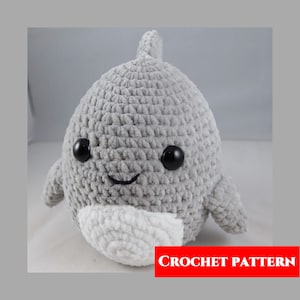 May include: A gray crocheted plush dolphin with black eyes and a white belly. The dolphin has a smiling expression. CROCHET PATTERN is written in red letters at the bottom of the image.
