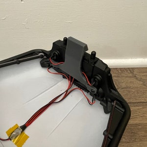 May include: A close-up of the inside of a black plastic toy car. The car has a gray plastic latch and red and black wires. The wires are connected to a yellow electrical tape.