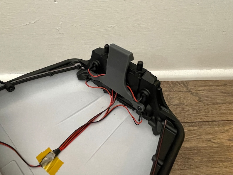 May include: A close-up of the inside of a black plastic toy car. The car has a gray plastic latch and red and black wires. The wires are connected to a yellow electrical tape.
