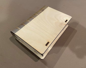Peut inclure: Une boîte en bois de couleur claire, conçue comme un livre. La boîte a un dos incurvé avec des lignes décoratives et de petits trous. La boîte rectangulaire a un couvercle à charnière et est en bois de couleur claire.