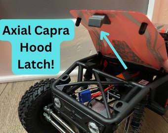 Op de afbeelding: Een rode Axial Capra RC-auto met de motorkap open, waardoor de motor en elektronica zichtbaar zijn. De motorkapvergrendeling is zichtbaar aan de bovenkant van de motorkap.