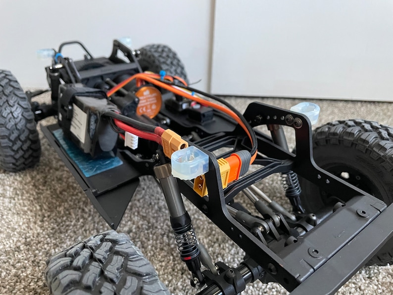 Pode incluir: Um chassi de carro RC preto com fios laranja e preto, uma bateria preta e p&aacute;ra-choques de pl&aacute;stico transparente. O carro est&aacute; em um tapete marrom.