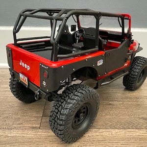 May include: Red Jeep Wrangler with a black roll cage and black wheels. The Jeep has a black rear bumper and a black spare tire carrier. The Jeep is on a wooden floor.