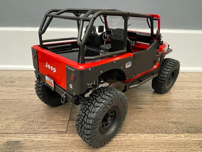 May include: Red Jeep Wrangler with a black roll cage and black wheels. The Jeep has a black rear bumper and a black spare tire carrier. The Jeep is on a wooden floor.