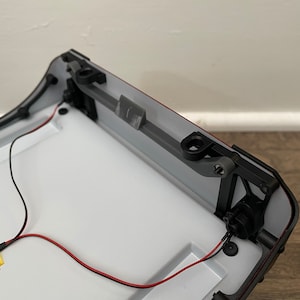 May include: A close-up of the underside of a white and black model car. The car has a red trim and a black plastic hinge. There are two black wires connected to the car.