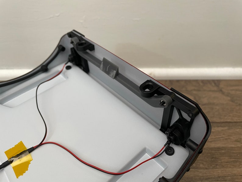 May include: A close-up of the underside of a white and black model car. The car has a red trim and a black plastic hinge. There are two black wires connected to the car.