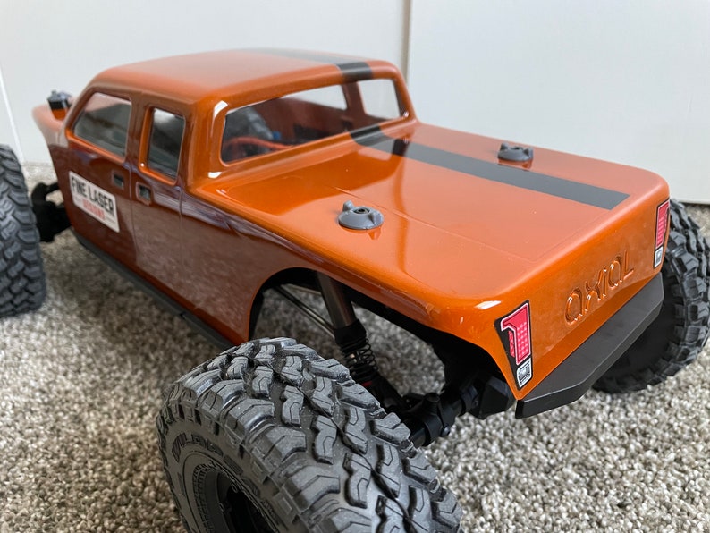 Pode incluir: Um close-up de um caminh&atilde;o de brinquedo marrom e preto com pneus pretos. O caminh&atilde;o tem uma faixa preta na ca&ccedil;amba e o texto "Axial" na parte de tr&aacute;s.