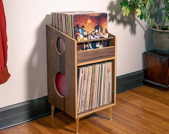 May include: A dark wood vinyl record storage stand with two tiers for holding albums. The side panels feature two circular cutouts. The stand is supported by four light-colored legs and contains a large collection of vinyl records in sleeves.