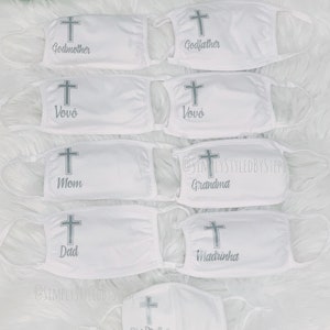 Op de afbeelding: Set witte gezichtsmaskers met een zilveren kruis en verschillende titels, waaronder "Peter", "Meter", "Vov&oacute;", "Moeder", "Vader", "Oma", "Peetmoeder" en "Grote broer".