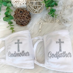 Op de afbeelding: Twee witte gezichtsmaskers met een zilveren glitterkruis en de woorden "Peetmoeder" en "Peter" erop gedrukt.