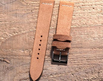 May include: A tan suede watch strap featuring a textured surface, white contrast stitching near the attachment points, six circular adjustment holes, two matching suede keepers, and a polished silver-tone metal buckle for secure fastening.