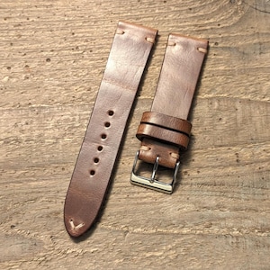 Può includere: Un cinturino per orologio in pelle marrone con cuciture chiare, una fibbia in metallo color argento e due passanti in pelle abbinati. Il cinturino presenta una serie di fori di regolazione circolari e un'estremità affusolata con cuciture rinforzate.