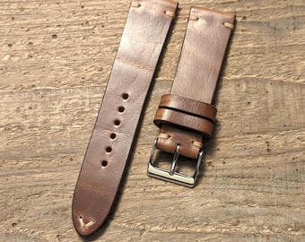May include: A brown leather watch strap featuring light-colored stitching, a silver-toned metal buckle, and two matching leather keepers. The strap includes a series of circular adjustment holes and a tapered end with reinforced stitching details.