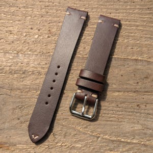 May include: A two-piece dark brown leather watch strap featuring light beige contrast stitching at the attachment points and tip. The strap includes a silver-toned metal buckle, two leather keepers, and six circular adjustment holes on the long end.