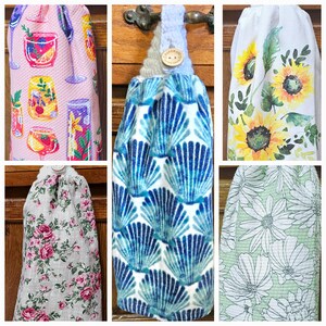 May include: Five different kitchen towels with various patterns. The towels are hanging on hooks. The patterns include a pink and white floral design, a blue and white seashell design, a white towel with a sunflower design, a green towel with a white daisy design, and a gray and white floral design.