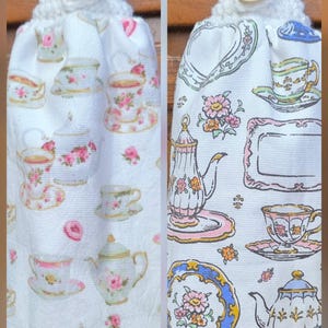 May include: A white hanging kitchen towel featuring a repeating pattern of colorful teapots, teacups, and floral saucers. The towel has a white crocheted top with a wooden button labeled handmade with love, designed to hang from a cabinet handle.