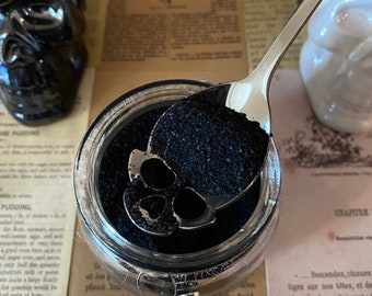 Puede incluir: Un tarro de vidrio lleno de polvo de carbón vegetal negro activado. Una cuchara de plata está sacando el carbón. El tarro está sentado en una mesa con un libro y una calavera decorativa.