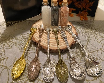 May include: A collection of decorative spoons in gold, silver, and bronze tones, with ornate floral and seashell designs. Three small glass bottles with cork stoppers sit on a wooden slice, filled with white, black, and pink contents.