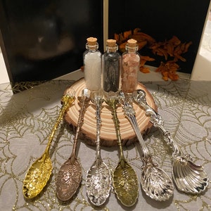 May include: A collection of decorative spoons in gold, silver, and bronze tones, with ornate floral and seashell designs. Three small glass bottles with cork stoppers sit on a wooden slice, filled with white, black, and pink contents.