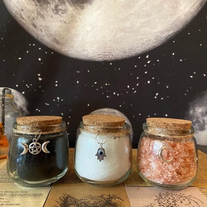 May include: Three glass jars with cork lids, filled with black powder, white crystals, and pink salt. Each jar has a silver charm: a triple moon and pentacle, a Hamsa hand, and a crescent moon. The background features a moon and stars.