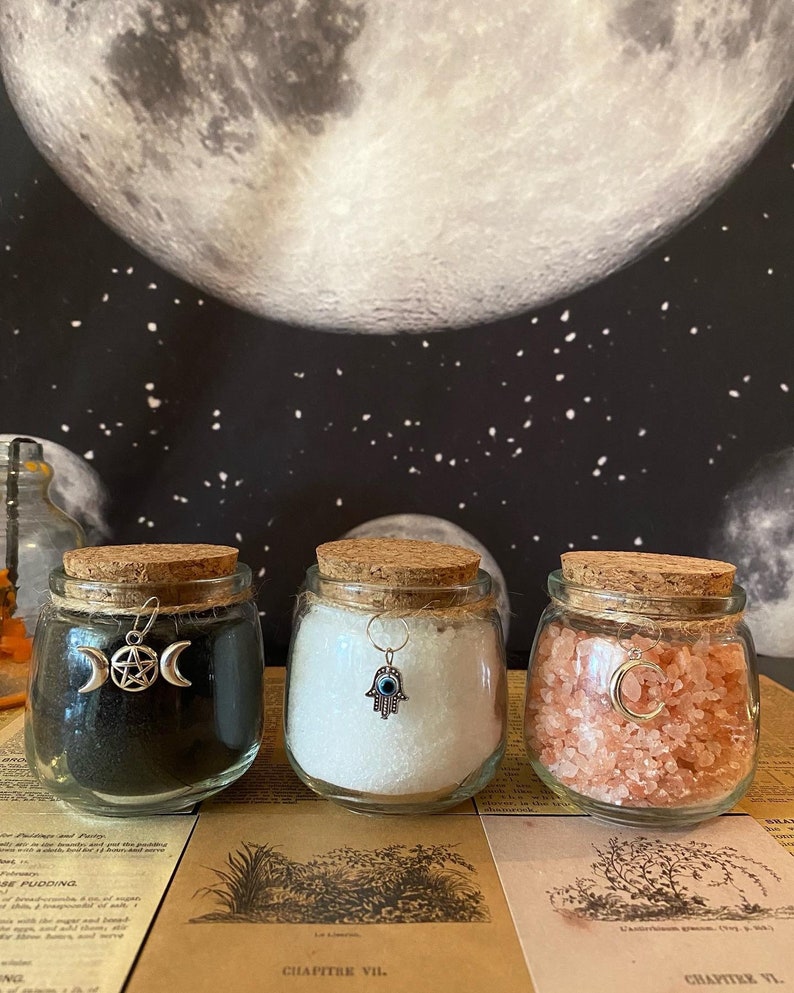 May include: Three glass jars with cork lids, filled with black powder, white crystals, and pink salt. Each jar has a silver charm: a triple moon and pentacle, a Hamsa hand, and a crescent moon. The background features a moon and stars.