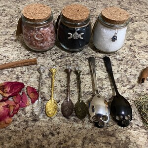 May include: Three glass jars with cork lids, filled with pink salt, black powder, and white salt, are displayed. Decorative spoons, rose petals, cinnamon sticks, and herbs are arranged around the jars. The jars have charms attached to them.