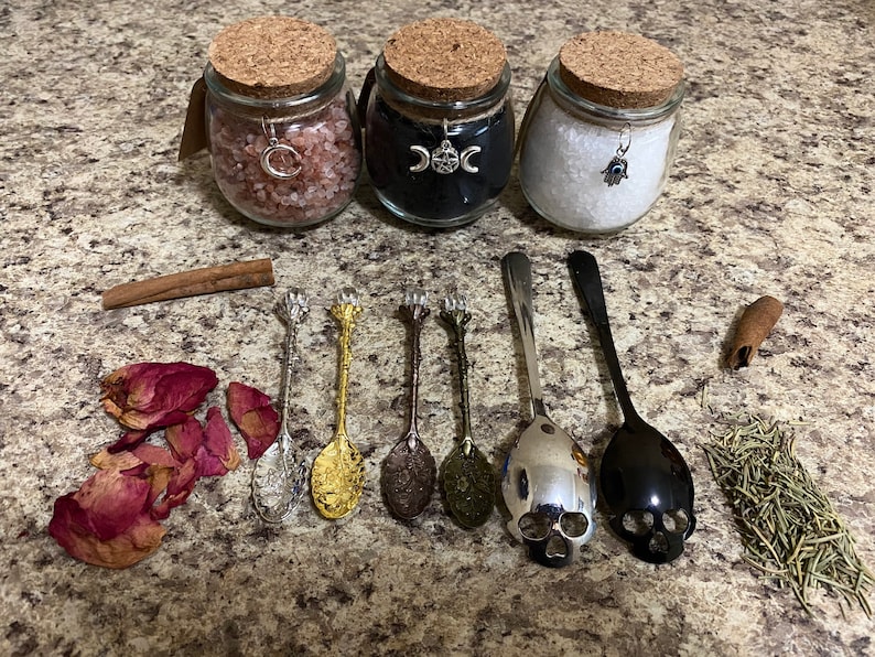 May include: Three glass jars with cork lids, filled with pink salt, black powder, and white salt, are displayed. Decorative spoons, rose petals, cinnamon sticks, and herbs are arranged around the jars. The jars have charms attached to them.