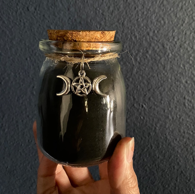 May include: A small, clear glass jar filled with black powder, sealed with a cork stopper and twine. A silver charm with a triple moon and pentacle design hangs from the jar. The jar is held against a dark blue background.