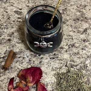 May include: A glass jar filled with black sand, adorned with a silver triple moon charm and twine. A gold-colored spoon with a crystal top rests inside. Dried rose petals, rosemary, and cinnamon sticks are arranged nearby.