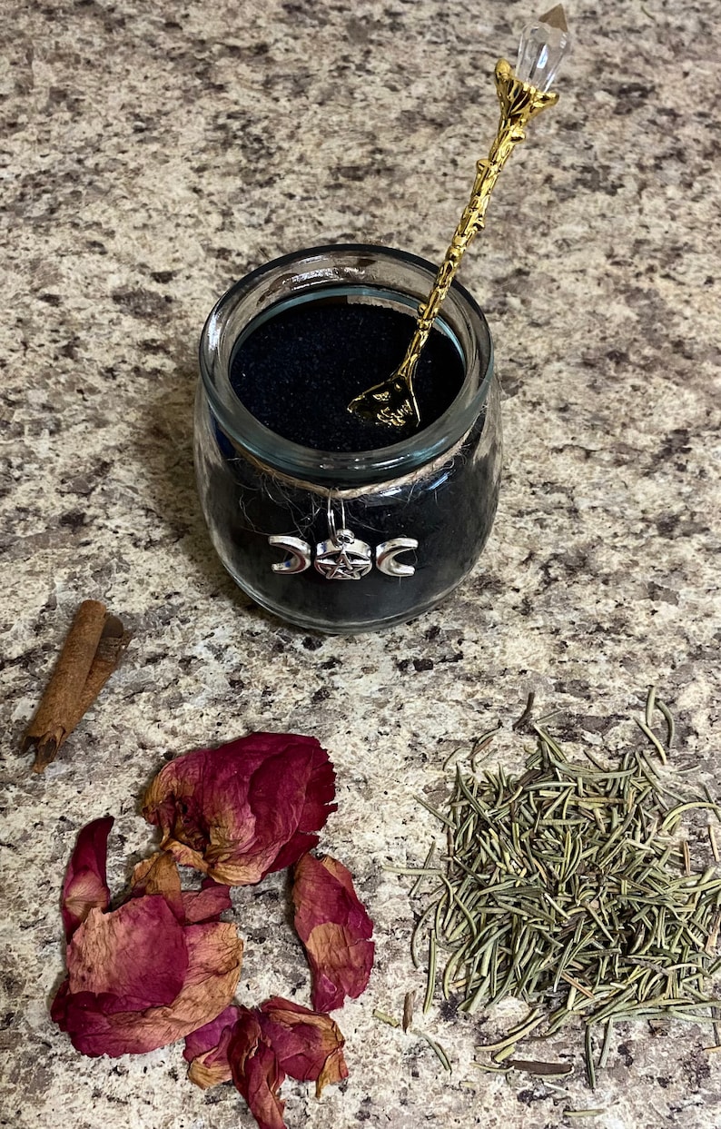 May include: A glass jar filled with black sand, adorned with a silver triple moon charm and twine. A gold-colored spoon with a crystal top rests inside. Dried rose petals, rosemary, and cinnamon sticks are arranged nearby.