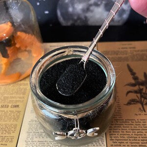 Puede incluir: Un tarro de vidrio lleno de arena volc&aacute;nica negra, una cuchara de plata con un alambre de cobre envuelto y un amuleto de plata con un s&iacute;mbolo de triple luna.