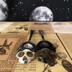 May include: Two spoons with skull-shaped bowls. One spoon is silver, the other is black. The spoons are placed on a vintage newspaper background, with a backdrop of a night sky with moons and stars. The spoons are a unique kitchen or dining accessory.
