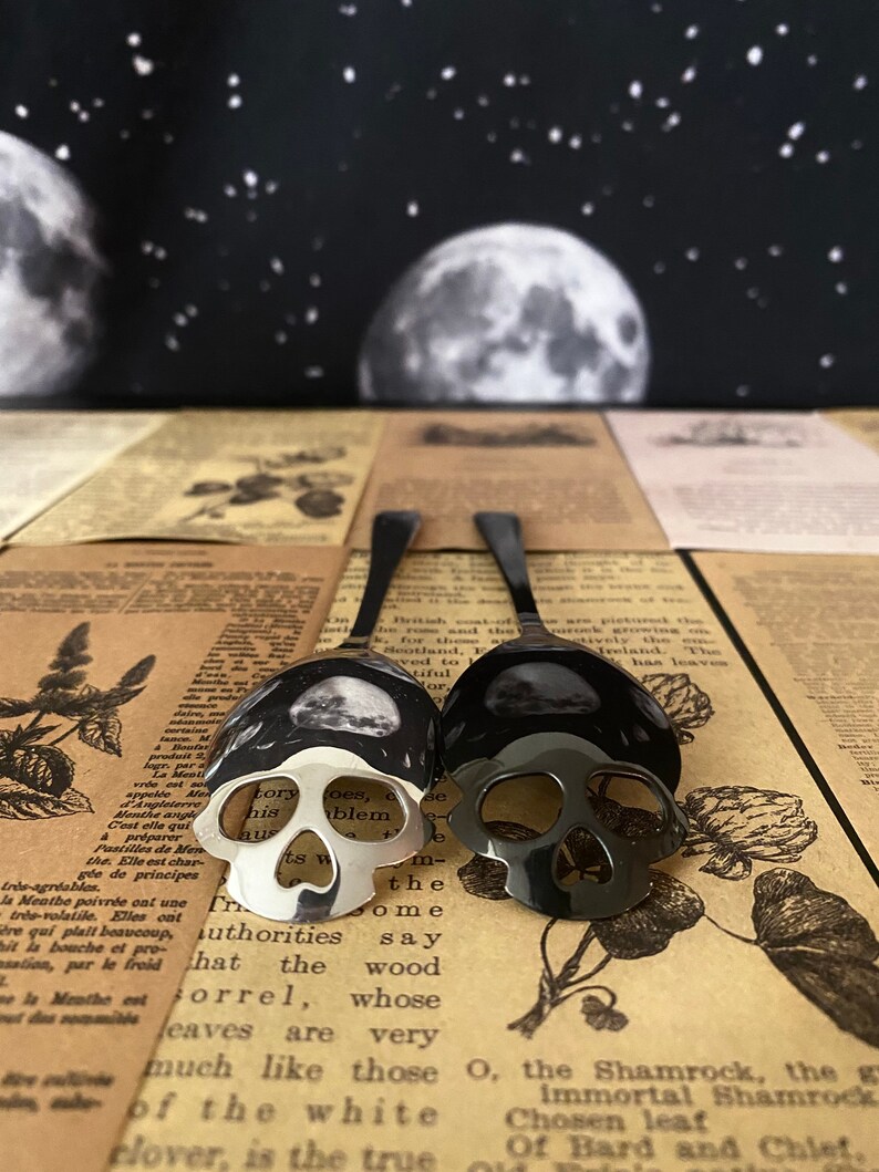 May include: Two spoons with skull-shaped bowls. One spoon is silver, the other is black. The spoons are placed on a vintage newspaper background, with a backdrop of a night sky with moons and stars. The spoons are a unique kitchen or dining accessory.
