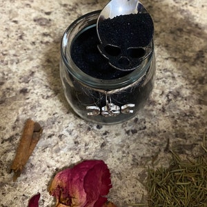 May include: A glass jar filled with black granules, a silver spoon with a skull-shaped cutout, and a decorative charm. The jar is adorned with twine and a charm featuring the triple moon symbol. Dried rose petals, rosemary, and a cinnamon stick are also present.