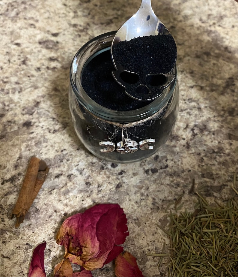 May include: A glass jar filled with black granules, a silver spoon with a skull-shaped cutout, and a decorative charm. The jar is adorned with twine and a charm featuring the triple moon symbol. Dried rose petals, rosemary, and a cinnamon stick are also present.