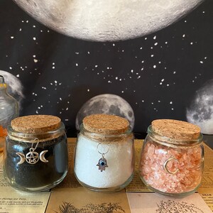 Puede incluir: Tres frascos de vidrio con tapas de corcho, cada uno lleno de un tipo diferente de sal. Los frascos est&aacute;n decorados con amuletos de plata: una luna creciente y un pentagrama, una mano de hamsa con un mal de ojo y una luna creciente.