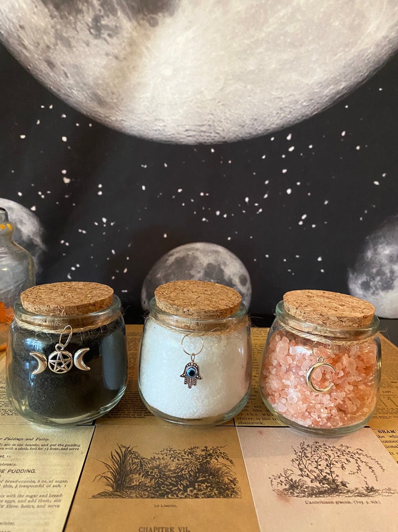 Puede incluir: Tres frascos de vidrio con tapas de corcho, cada uno lleno de un tipo diferente de sal. Los frascos est&aacute;n decorados con amuletos de plata: una luna creciente y un pentagrama, una mano de hamsa con un mal de ojo y una luna creciente.