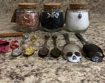 May include: Three glass jars with cork lids, filled with pink, black, and white contents, each adorned with a charm. Six decorative spoons with ornate handles and skull-shaped scoops are arranged in front. A cinnamon stick and rose petals are also present.