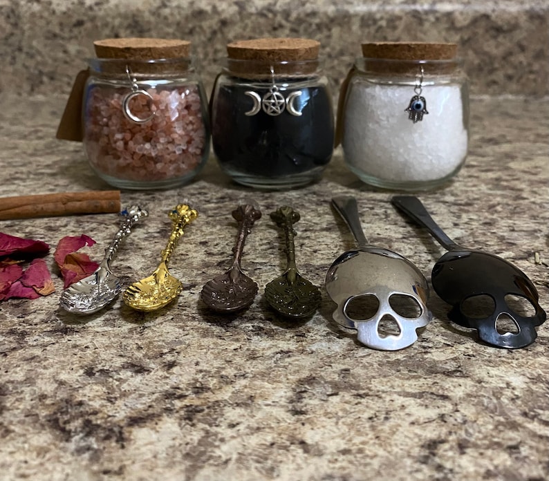 May include: Three glass jars with cork lids, filled with pink, black, and white contents, each adorned with a charm. Six decorative spoons with ornate handles and skull-shaped scoops are arranged in front. A cinnamon stick and rose petals are also present.
