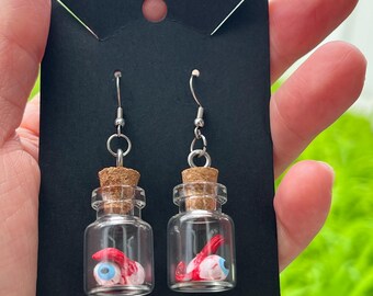 Puede incluir: Un par de pendientes de fantasía con botellas de vidrio en miniatura con tapones de corcho. Cada botella contiene un pequeño globo ocular detallado con detalles en rojo. Los pendientes cuelgan de ganchos plateados, presentados en una tarjeta negra.